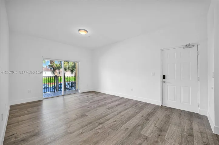 Open-concept interior featuring wood-finish flooring, white walls, flush-mount ceiling lighting, a multi-panel entry door, and sliding glass doors to a balcony with metal railing