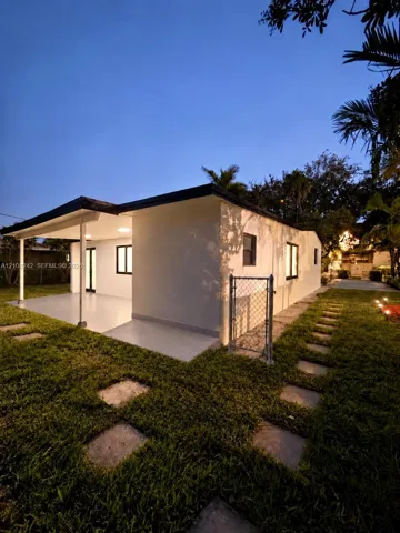 Exterior elevation featuring a light-toned smooth stucco finish, a covered patio with tiled flooring, stone paver walkways, and a chain-link fence enclosure