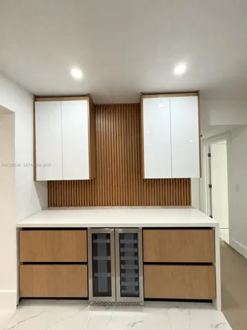 Custom built-in cabinetry featuring white gloss upper cabinets, wood-finish lower drawers, integrated wine refrigerator, white stone-surface countertop, and vertical wood slat backsplash