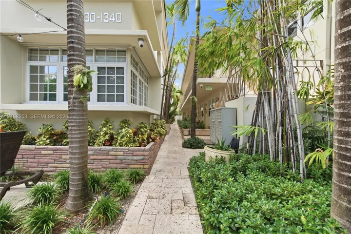 Exterior walkway featuring stone paver flooring, tiered stone-finish planters, multi-pane windows, and lush tropical landscaping