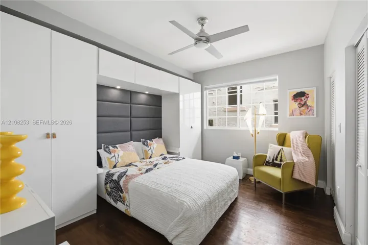 Room featuring dark wood-finish flooring, built-in white cabinetry, integrated recessed lighting, a charcoal grey upholstered headboard, and a multi-blade ceiling fan