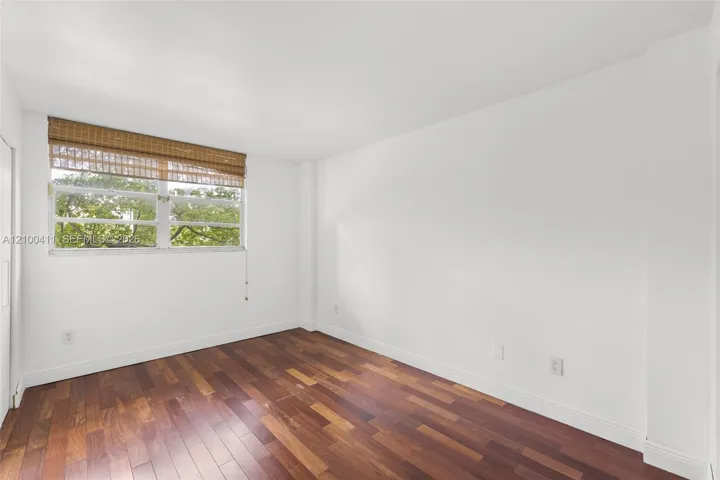Interior space featuring dark wood-finish flooring, crisp white walls, and a multi-pane window with a woven-texture shade