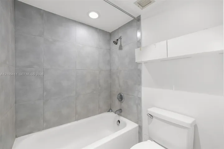 Full-height grey tile tub surround, white porcelain bathtub, wall-mounted shower head, recessed ceiling lighting, and built-in white upper cabinetry