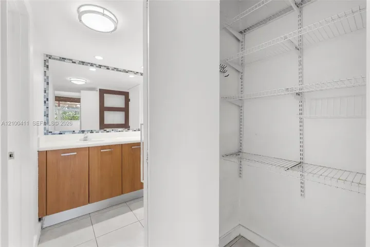 Bathroom featuring a wood-finish vanity with a solid-surface countertop, tiled flooring, a framed mirror with a decorative mosaic border, and a walk-in storage area with adjustable wire shelving