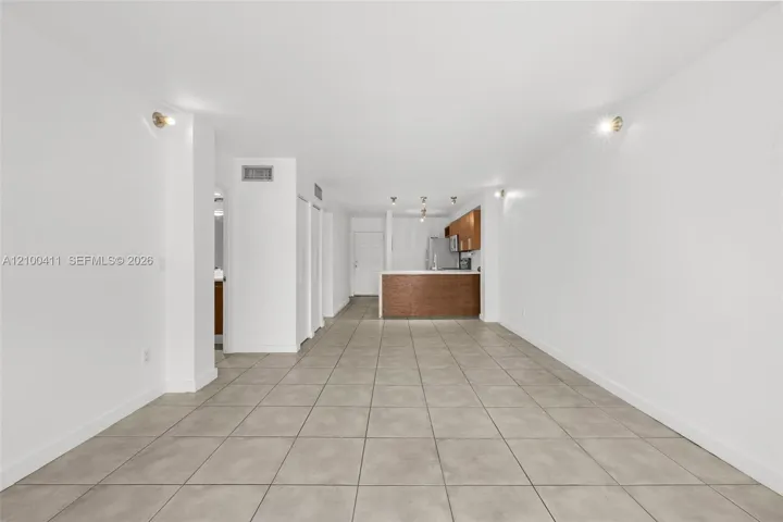 Neutral-toned tile flooring throughout the open-concept interior, featuring wall-mounted lighting fixtures, a kitchen peninsula with wood-finish cabinetry, and recessed lighting