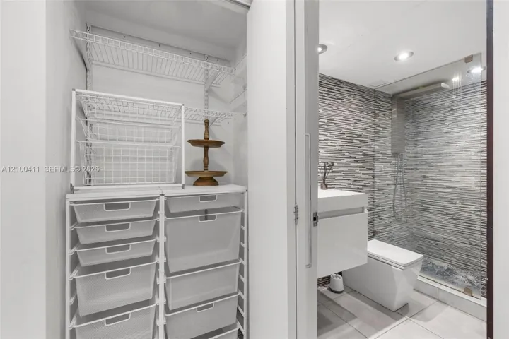 Walk-in closet featuring wire shelving and a modular storage system, paired with a bathroom containing a wall-mounted vanity, square-profile toilet, glass-enclosed shower with horizontal textured tile, and recessed lighting