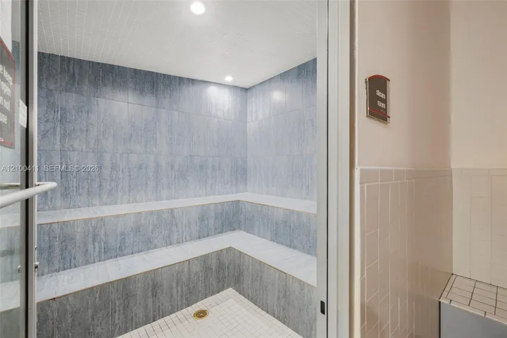 Steam room featuring full-height blue-toned tile wall cladding, tiered built-in bench seating, recessed lighting, and a white tiled floor