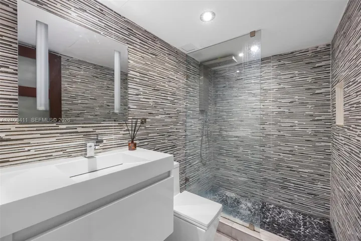 Modern floating vanity with a seamless integrated sink, wall-mounted faucet, linear multi-toned tile wall covering, frameless glass shower enclosure, and recessed shower shelving