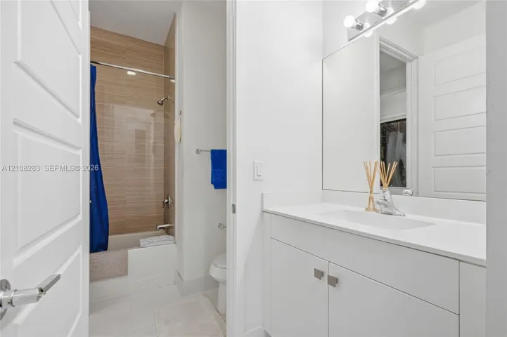 Bathroom featuring a white vanity with a composite countertop, an integrated rectangular sink, a floor-to-ceiling tiled shower surround with a light wood-finish pattern, and a wall-mounted light fixture above a large mirror