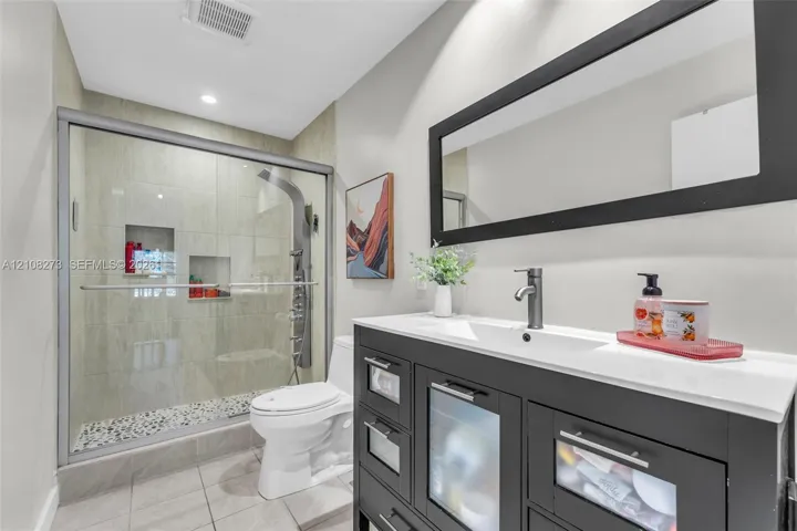 Dark-finish vanity with white countertop and rectangular integrated sink, walk-in shower with stone tile flooring and recessed shelving, and a large rectangular mirror with a black frame