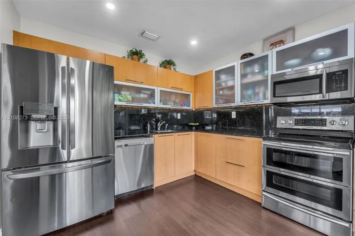 Kitchen featuring light wood-finish cabinetry with glass-front uppers, dark stone countertops, stainless steel appliances, and recessed ceiling lighting