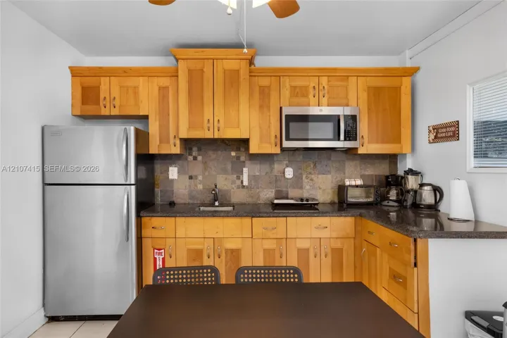 Kitchen featuring natural wood cabinetry, stone-tile backsplash, dark stone countertops, stainless steel refrigerator, and built-in microwave