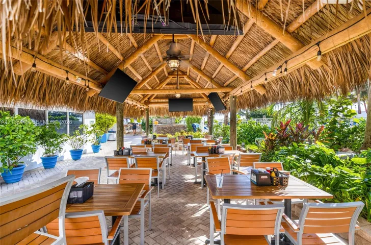 Covered outdoor pavilion featuring a thatched-style roof, exposed timber framing, a central ceiling fan, paver flooring, and integrated string lighting
