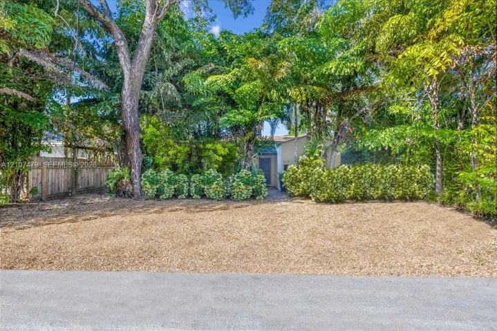Gravel-surfaced exterior area featuring mature trees, manicured hedges, and a vertical-plank wood fence