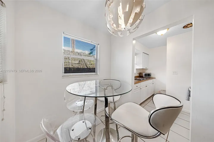 Dining area featuring a modern sculptural pendant light fixture, ceramic tile flooring, and a window with blinds, opening into a kitchen equipped with white cabinetry and stone countertops