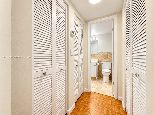 Hallway featuring parquet wood-finish flooring, multiple sets of white louvered-door closets, and a doorway entry to a tiled bathroom
