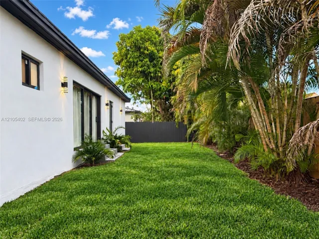 Exterior featuring white stucco siding, black-framed glass sliding doors, dark metal wall-mounted exterior lighting, manicured lawn, and mature palm trees enclosed by a charcoal-colored privacy fence