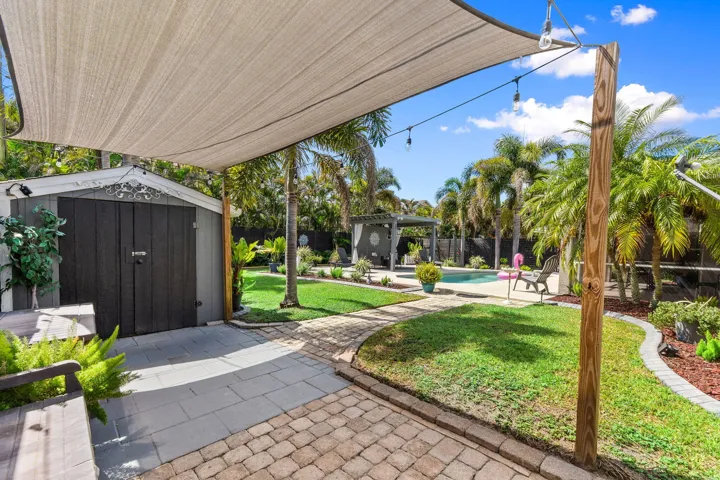 Outdoor living area featuring a shade sail, paved stone patio, swimming pool, detached storage structure, and established palm trees