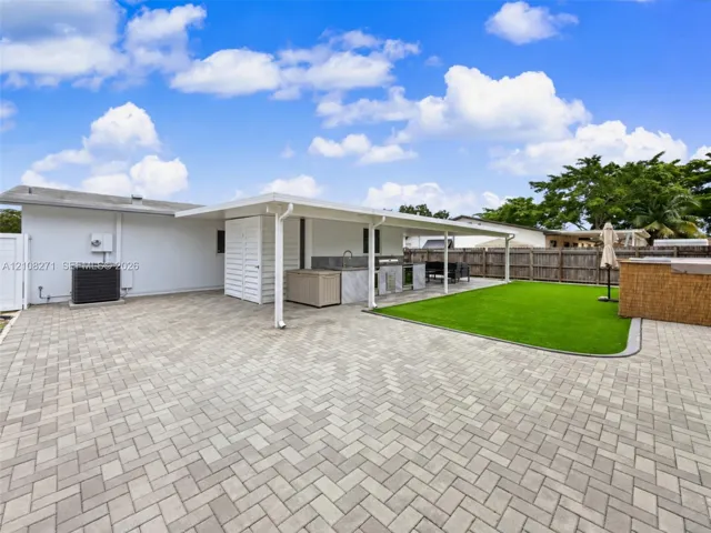 Rear exterior featuring a covered patio, paved hardscaping, synthetic turf area, and built-in outdoor kitchen with stone-finish cabinetry
