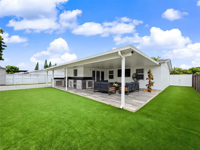 Covered patio featuring wood-finish tile flooring, integrated outdoor kitchen with stone veneer and dark countertop, and synthetic turf lawn