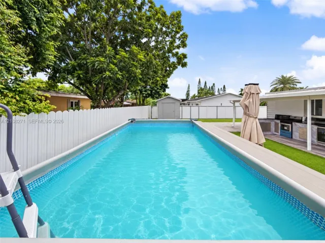 Rectangular swimming pool featuring a tiled waterline, composite decking surround, built-in outdoor kitchen with stone-finish cabinetry, and a white privacy fence