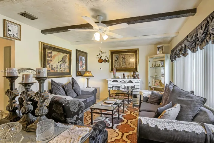 Living area featuring exposed ceiling beams, a white ceiling fan with integrated lighting, and a brick-faced fireplace