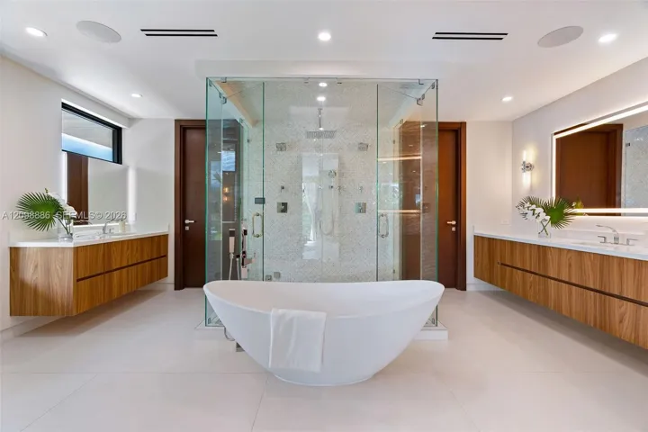 Dual floating wood-finish vanities with rectangular vessel sinks, floor-to-ceiling glass-enclosed shower with tiled interior, and freestanding soaking tub