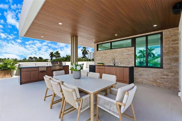 Covered outdoor living space featuring a wood-finish ceiling with recessed lighting, stone veneer wall, integrated kitchenette with stainless steel grill, cabinetry with dark countertops, and large-format tile flooring