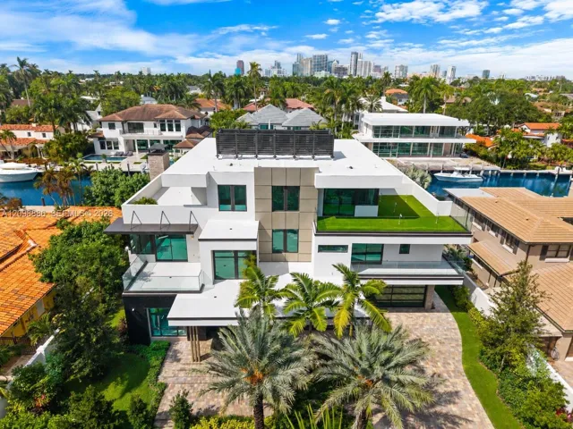 Multi-level contemporary residence featuring a flat roofline, glass panel balcony railings, stone-paved driveway, elevated terrace with turf surfacing, and waterfront placement