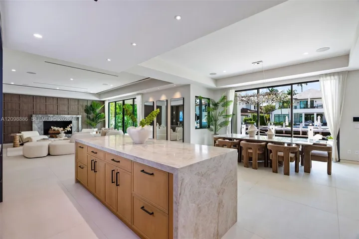 Large kitchen island featuring a waterfall stone countertop and light wood-finish cabinetry with matte black hardware, stone tile flooring, a floor-to-ceiling stone fireplace surround, and expansive sliding glass doors
