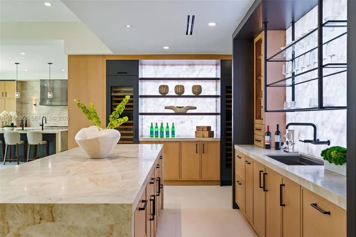 Custom kitchen featuring a large stone-surfaced island, light wood cabinetry, integrated wine refrigeration, an illuminated stone backsplash, and matte black hardware