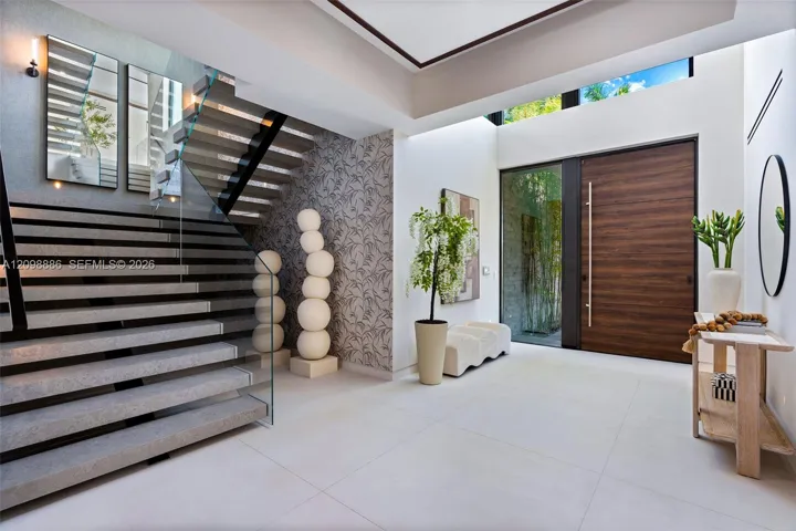 Grand foyer featuring large-format tile flooring, a custom wood-finished entry door with a vertical pull handle, a dramatic staircase with glass railings, and a double-height ceiling with clerestory windows