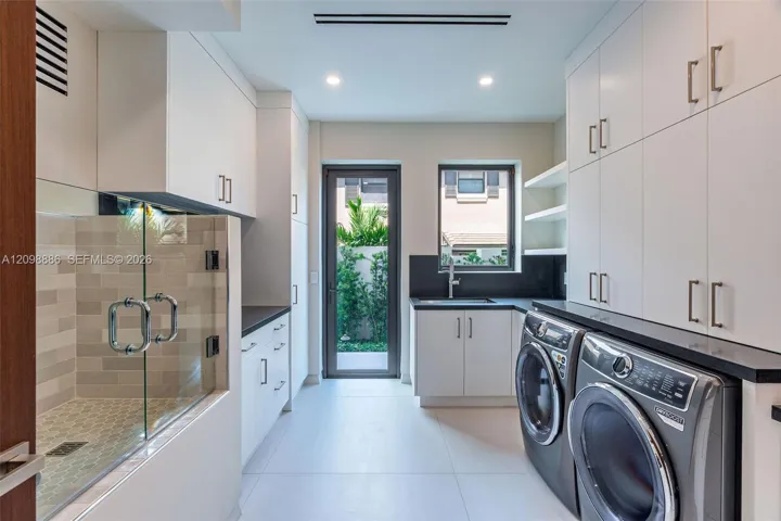 Utility room featuring extensive white cabinetry, dark solid-surface countertops, a deep undermount sink, porcelain tile flooring, and an enclosed tiled shower