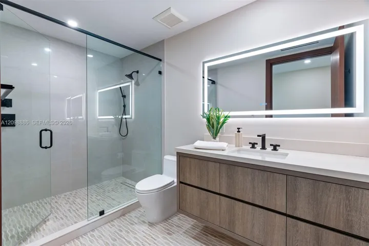 Modern bathroom featuring a wood-finish floating vanity with a white countertop, walk-in glass-enclosed shower with matte black fixtures, and a large backlit wall-mounted mirror
