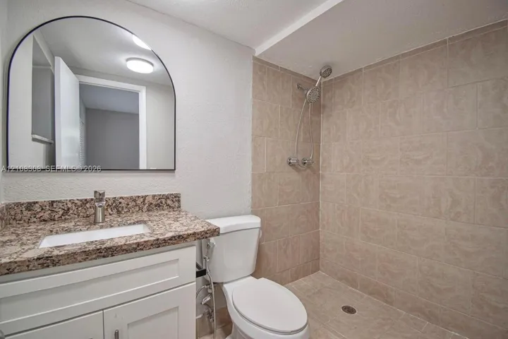 Bathroom featuring a white vanity with a stone-finish countertop, undermount sink, arched wall mirror, and walk-in shower with neutral tile surround and adjustable handheld showerhead