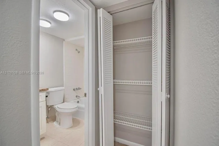 Built-in storage closet with louvered bi-fold doors and wire shelving, featuring a tiled bathtub enclosure, porcelain toilet, and recessed ceiling lighting