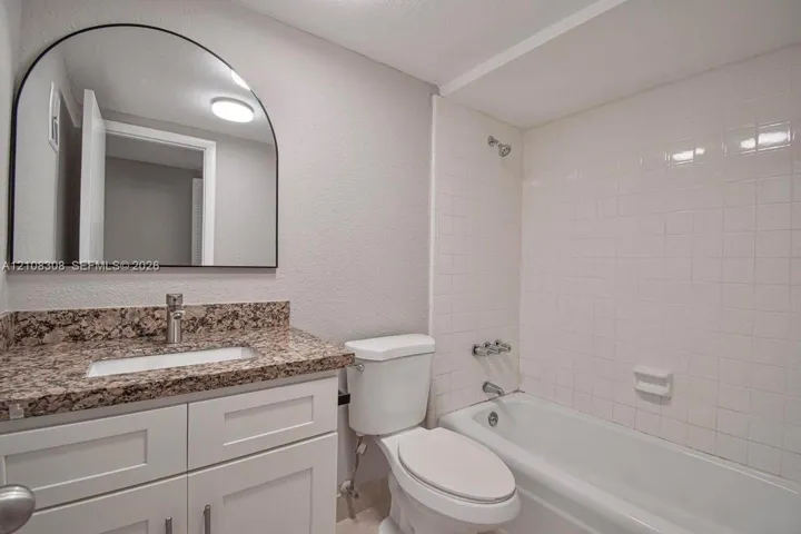 White cabinetry with stone-finish countertop, arched wall mirror, and tiled shower-tub combination
