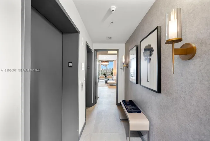 Hallway featuring textured grey wall cladding, gold-toned metallic wall sconces with cylindrical glass shades, and light-toned tile flooring