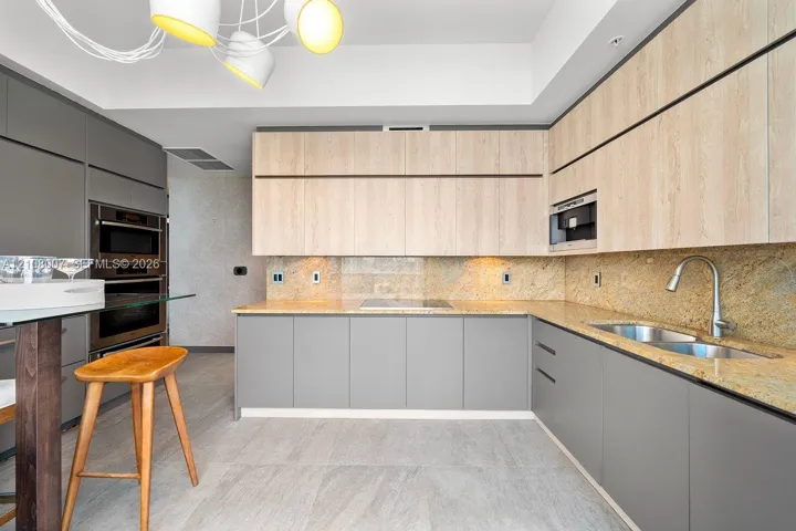 Kitchen featuring two-tone flat-panel cabinetry, stone countertops, integrated wall ovens, a multi-head modern light fixture, and stone-finish flooring