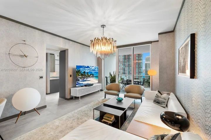 Living area featuring textured wall coverings, multi-tiered glass crystal chandelier, large-format tile flooring, and floor-to-ceiling glass sliding doors opening to an exterior balcony
