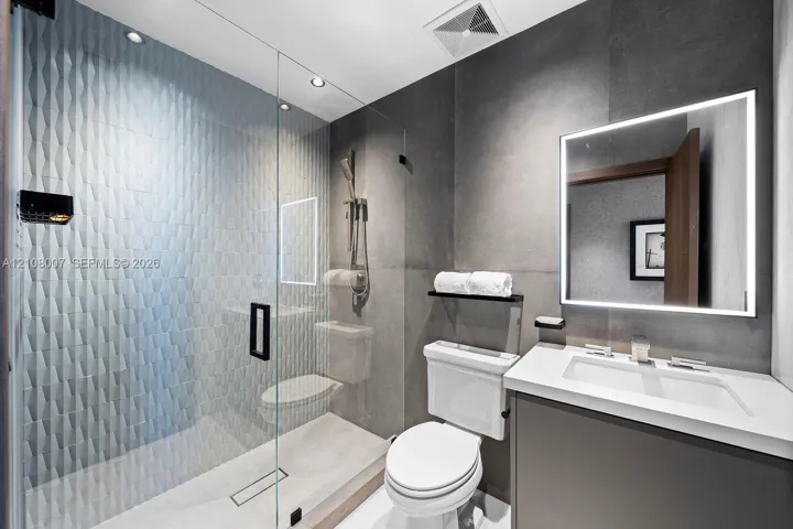 Walk-in glass-enclosed shower featuring three-dimensional tile accent wall, vanity with solid surface countertop, integrated LED-backlit mirror, and matte black fixtures