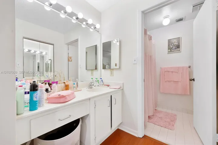 Full-width wall mirror, vanity featuring an integrated basin, wood-finish flooring, and multi-bulb Hollywood-style light fixture