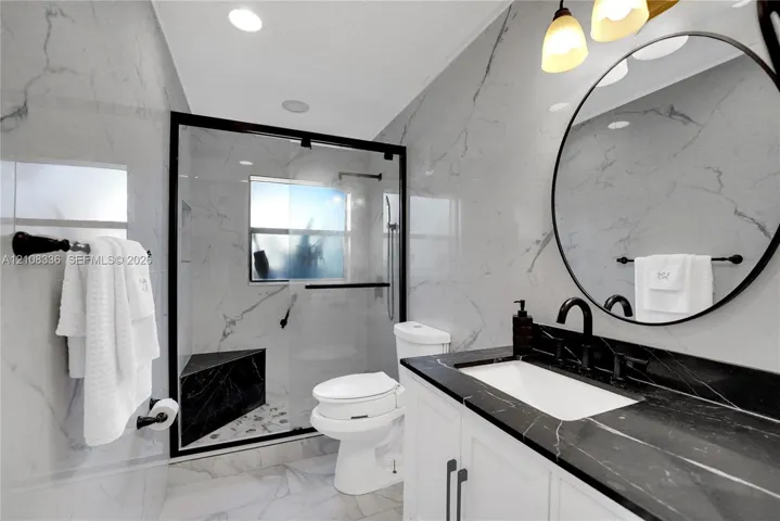 Bathroom featuring large-format wall tiling, a walk-in glass-enclosed shower with a built-in bench, a custom vanity with a contrasting dark stone countertop, a circular wall-mounted mirror, and matte black hardware