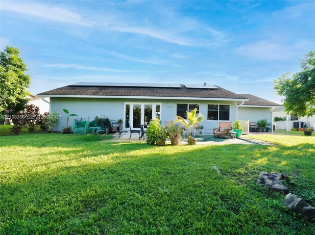 Exterior rear elevation featuring a stucco facade, rooftop solar panel array, multi-panel glass door, and a paved patio area set within a spacious lawn
