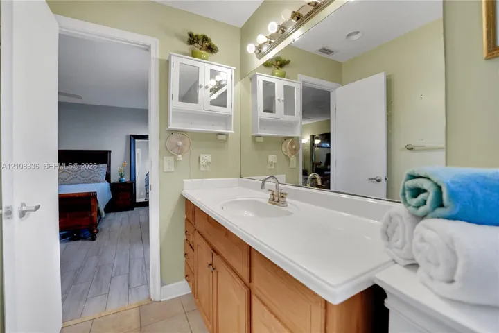 Single-sink vanity with a solid-surface countertop, natural wood-finish cabinetry, wall-mounted white storage hutch, and large-scale vanity mirror with integrated multi-bulb lighting