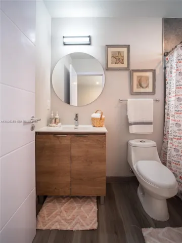 Bathroom featuring a wood-finish vanity with a rectangular sink, circular mirror, wall-mounted light fixture, and wood-finish flooring
