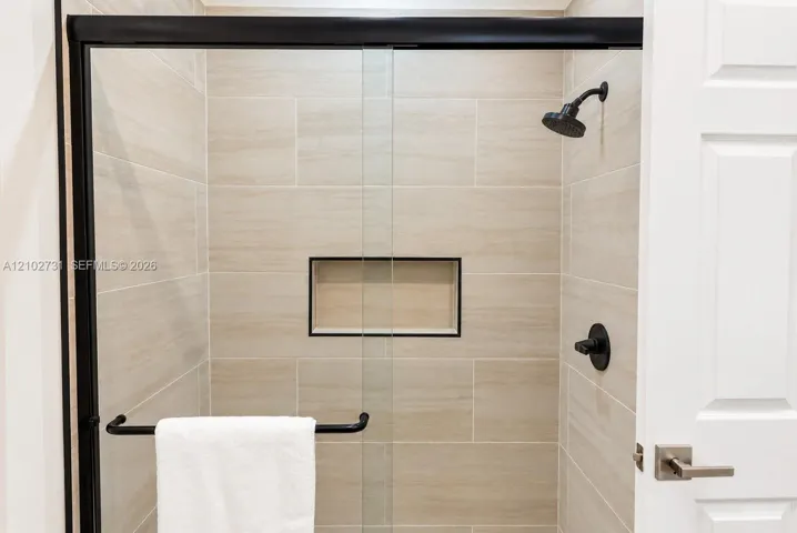 Walk-in shower featuring floor-to-ceiling neutral tile, a built-in recessed storage niche, a glass enclosure with matte black framing, and a matte black rainfall showerhead