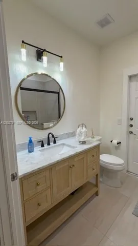 Bathroom vanity featuring light wood cabinetry, stone-finish countertop with an undermount sink, circular gold-framed mirror, three-light matte black fixture, and matte black hardware