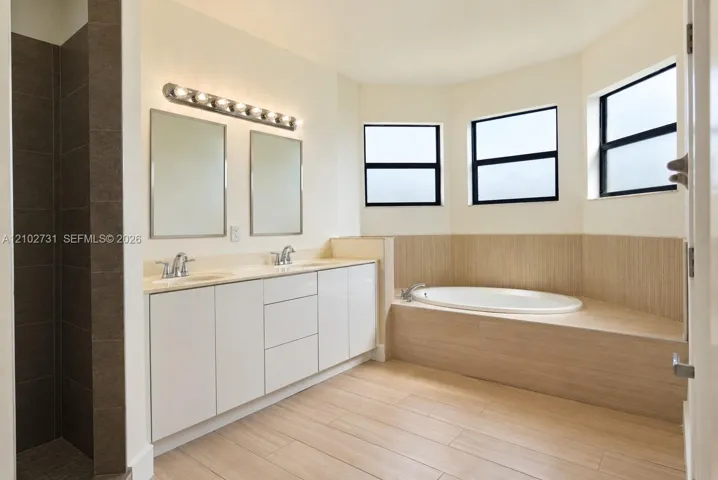 Bathroom featuring a dual-sink vanity with white cabinetry, a built-in soaking tub with tile surround, wood-finish flooring, and bay windows