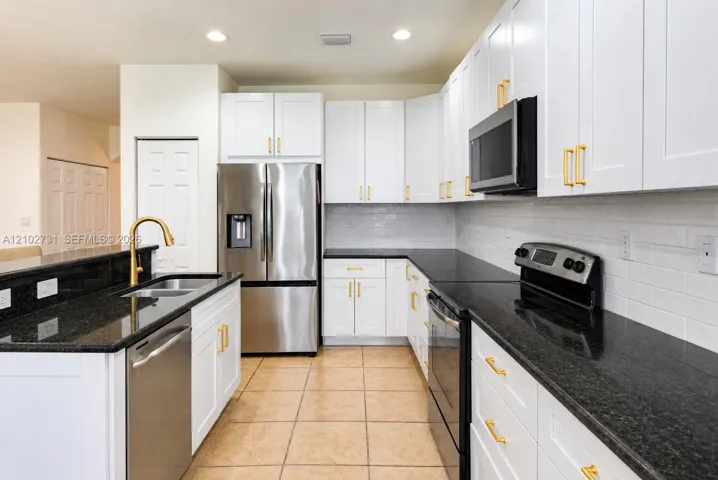 Kitchen featuring white shaker-style cabinetry, dark stone countertops, brass-finish hardware, stainless steel appliances, and a tiled backsplash